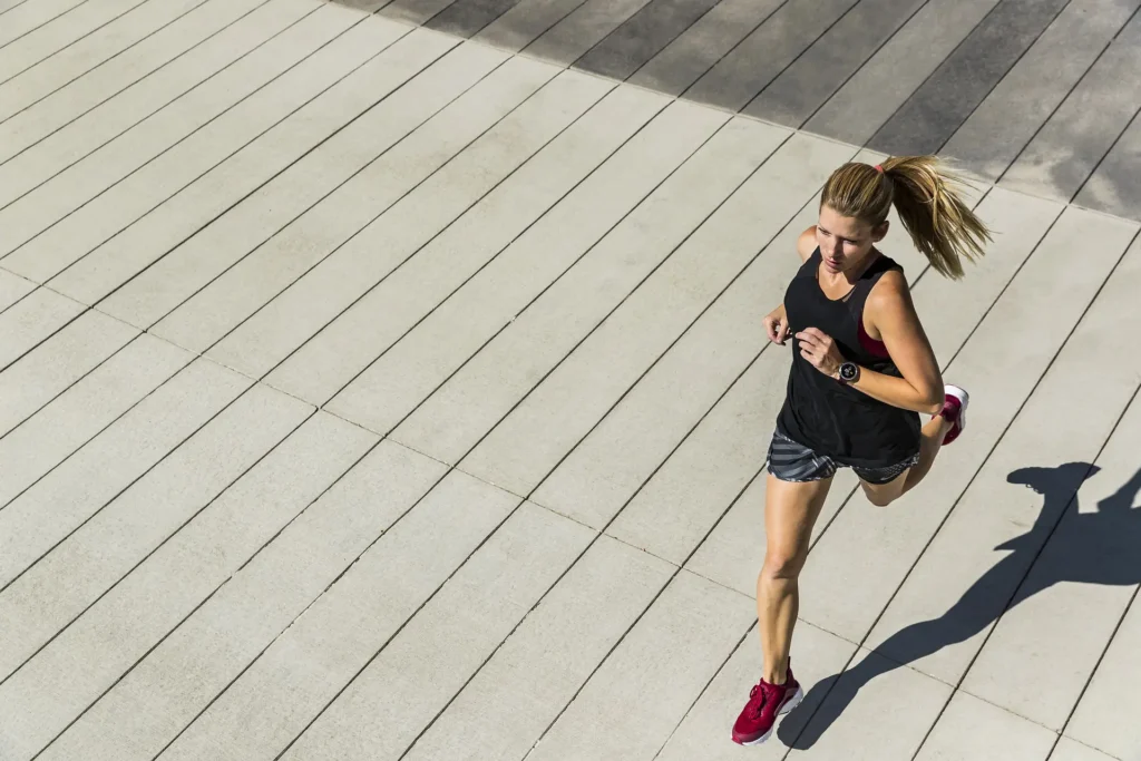 Persona che corre su un’ampia superficie pavimentata in lastre chiare, ripresa dall’alto mentre avanza con passo deciso. Indossa canotta e pantaloncini sportivi scuri, scarpe da running rosse e un orologio sportivo al polso. La luce del sole proietta un’ombra netta sul terreno, accentuando il dinamismo dell’azione.